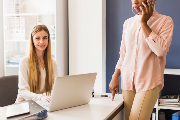 Mujer joven que usa la computadora portátil cerca de colega de la cosecha con smartphone