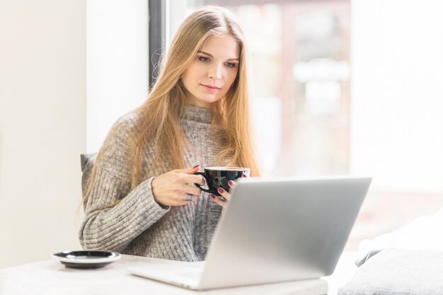 Mujer joven que usa la computadora portátil que se sienta con la taza