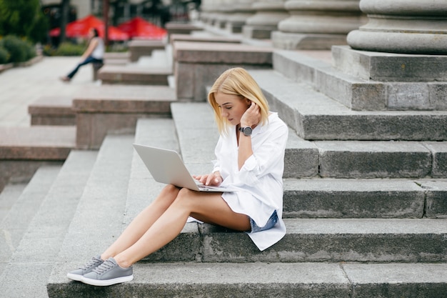 Mujer joven que usa la computadora portátil y el teléfono elegante. Muchacha hermosa del estudiante que trabaja en la computadora portátil al aire libre