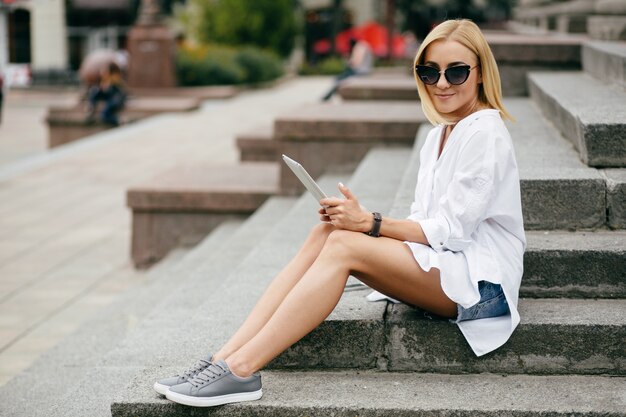 Mujer joven que usa la computadora portátil y el teléfono elegante. Muchacha hermosa del estudiante que trabaja en la computadora portátil al aire libre