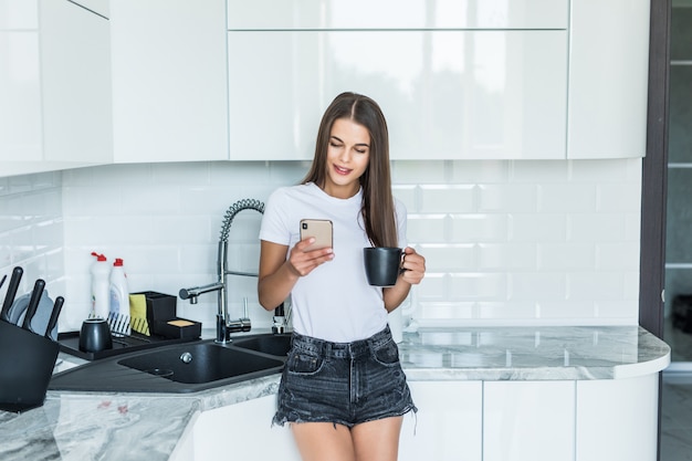 Mujer joven que usa el teléfono inteligente que se inclina en la mesa de la cocina con la taza de café en un hogar moderno. Mujer sonriente que lee el mensaje de teléfono. Chica morena feliz escribiendo un mensaje de texto