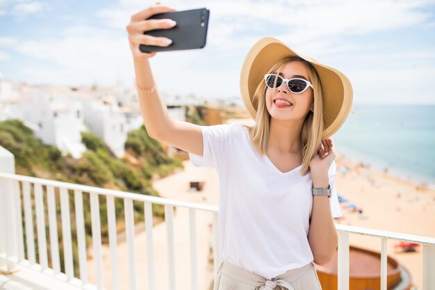 Mujer joven que viaja sosteniendo el teléfono haciendo selfie contra el mar