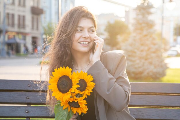 Una mujer joven con un ramo de girasoles está hablando por teléfono al atardecer