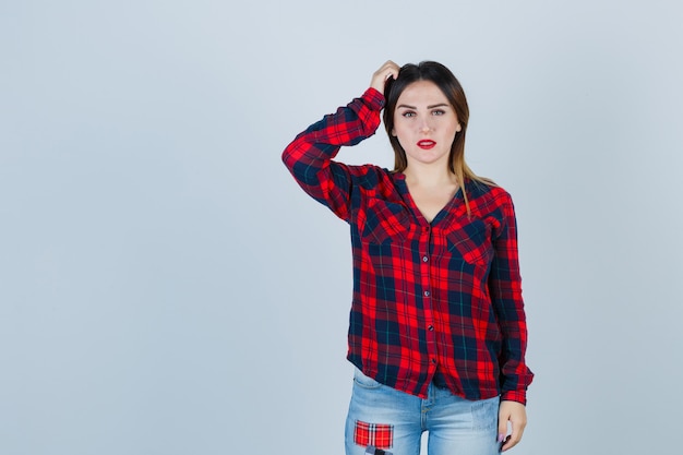 Mujer joven rascándose la cabeza en camisa a cuadros, jeans y mirando pensativo, vista frontal.