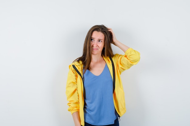 Mujer joven rascándose la cabeza en camiseta, chaqueta y mirando pensativo, vista frontal.