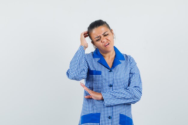 Mujer joven rascándose la cabeza mientras está parado en pose de pensamiento en camisa de pijama de cuadros azules y mirando pensativo. vista frontal.