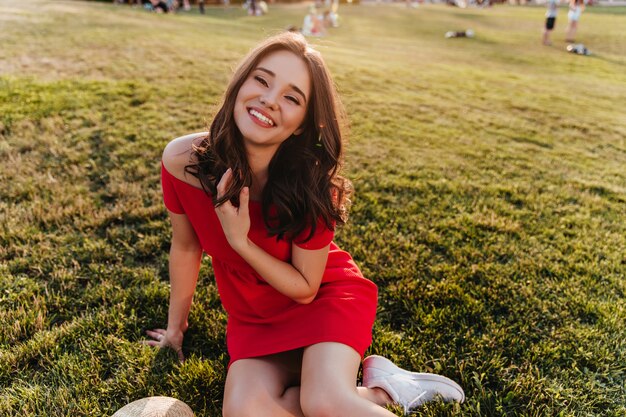 Mujer joven refinada en vestido rojo sentada en la hierba. Alegre niña sonriente con cabello oscuro posando sobre césped.