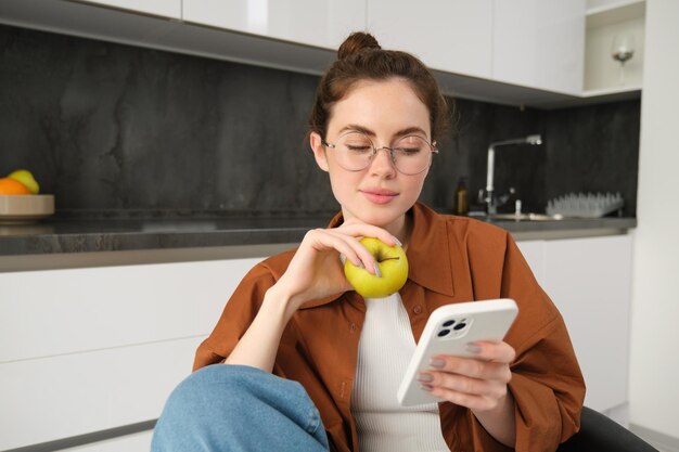 Mujer joven relajada en casa mirando su teléfono inteligente leyendo noticias en las redes sociales y comiendo verde