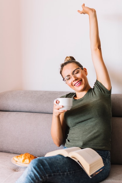 Foto gratuita mujer joven relajada feliz con el libro en el revestimiento que estira su mano que sostiene la taza de café