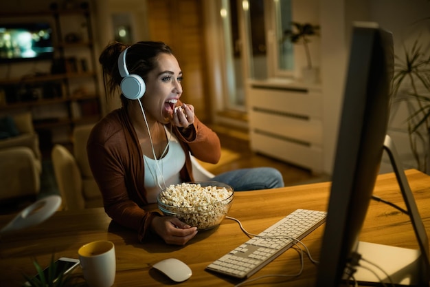 Mujer joven relajándose por la noche mientras ve una película en una computadora y come palomitas de maíz en casa