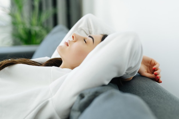 Mujer joven relajándose sentada en un sofá con las manos en la cabeza y mirando al aire libre a través de la ventana de la sala de estar en casa