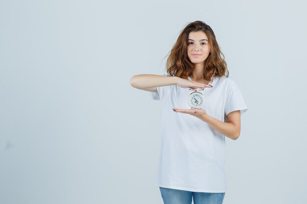 Mujer joven con reloj despertador en camiseta blanca, jeans y aspecto alegre. vista frontal.