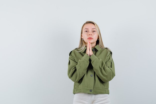 Mujer joven rezando en chaqueta verde, jeans y mirando impaciente, vista frontal.