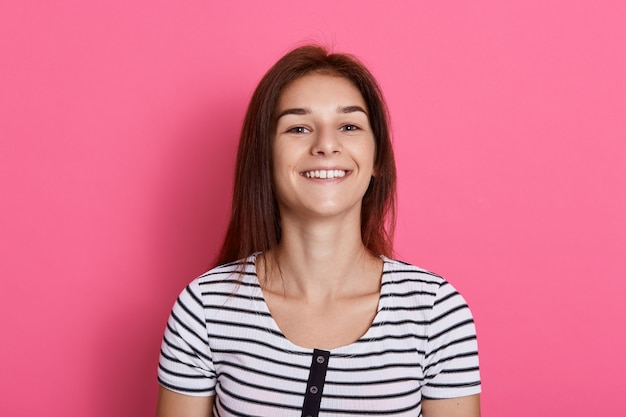 Mujer joven riendo contra la pared rosa, posando aislada sobre pared rosa, vistiendo camiseta a rayas, chica expresando emociones positivas.