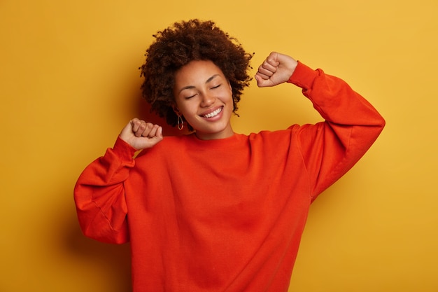 La mujer joven rizada alegre relajada sonríe feliz, mantiene ambas manos levantadas, lleva un jersey casual, tiene los ojos cerrados, modelos contra el fondo amarillo