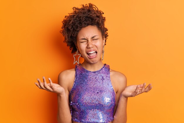 Mujer joven rizada disgustada extiende las palmas de las manos siente gritos frustrados ya que no ve salida viste ropa morada de moda hace que el peinado tenga un día desafortunado aislado sobre una pared naranja