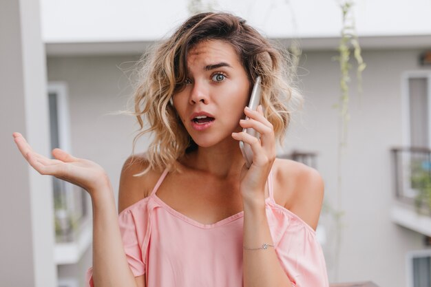 Mujer joven rizada preocupada en blusa rosa posando durante la conversación telefónica. Infeliz niña caucásica con cabello rubio con smartphone en el balcón.