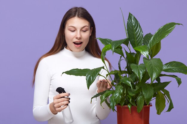 Mujer joven rocía plantas en macetas y mirando flores en maceta con la boca abierta