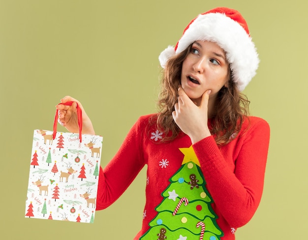 Mujer joven en rojo suéter de Navidad y gorro de Papá Noel con bolsa de papel con regalos de Navidad mirando a un lado desconcertado parado sobre pared verde