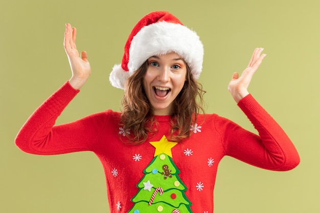 Mujer joven en rojo suéter de Navidad y gorro de Papá Noel feliz y emocionado levantando los brazos sobre fondo verde