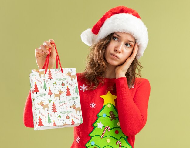 Mujer joven en rojo suéter de Navidad y gorro de Papá Noel sosteniendo una bolsa de papel con regalo de Navidad mirando a un lado desconcertado parado sobre la pared verde