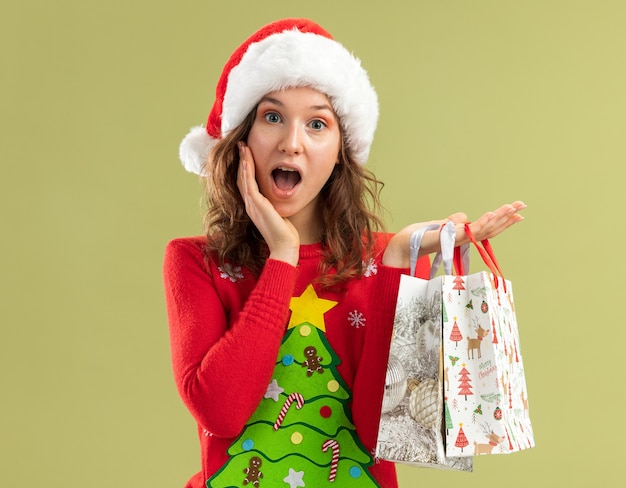 Mujer joven en rojo suéter de Navidad y gorro de Papá Noel sosteniendo bolsas de papel con regalos de Navidad feliz y asombrado de pie sobre la pared verde