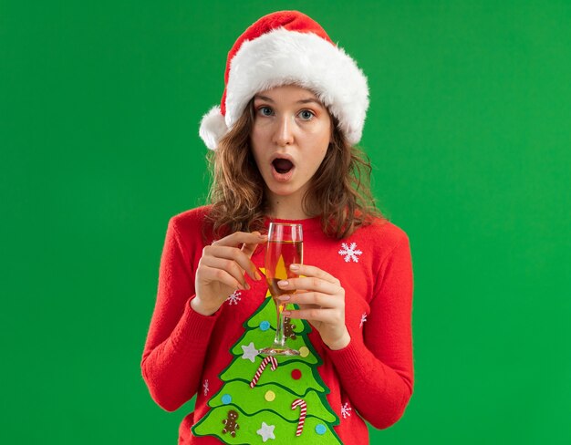 Mujer joven en rojo suéter de Navidad y gorro de Papá Noel sosteniendo una copa de champán mirando a la cámara asombrado y sorprendido de pie sobre fondo verde