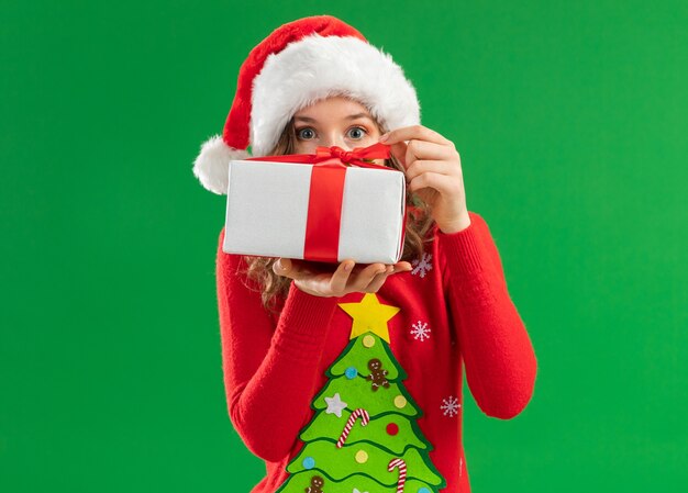 Mujer joven en rojo suéter de Navidad y gorro de Papá Noel sosteniendo un presente va a abrirlo intrigado de pie sobre fondo verde