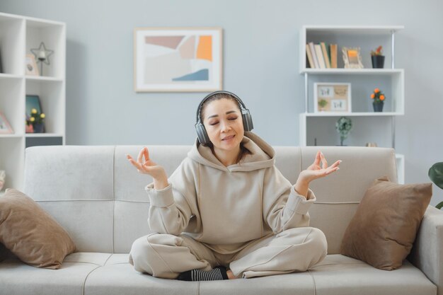 Mujer joven en ropa de casa sentada en un sofá en el interior de casa con auriculares feliz y positivo relajante meditando haciendo gesto de meditación con los dedos