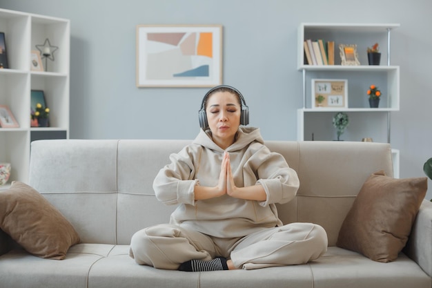 Mujer joven en ropa de casa sentada en un sofá en el interior de la casa con auriculares feliz y positivo relajante meditando haciendo gesto de meditación sosteniendo las palmas juntas