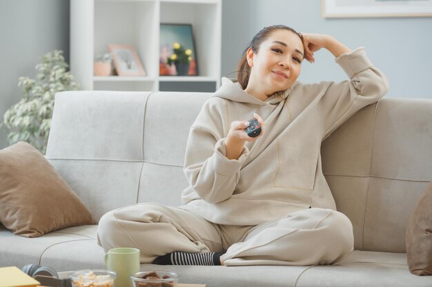 Mujer joven con ropa de casa sentada en un sofá en el interior de la casa sosteniendo un control remoto viendo la televisión con una sonrisa en la cara feliz y positiva