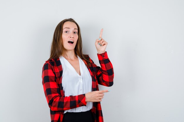 Mujer joven en ropa casual apuntando hacia arriba y hacia la derecha, mostrando un gesto de eureka y mirando inteligente, vista frontal.