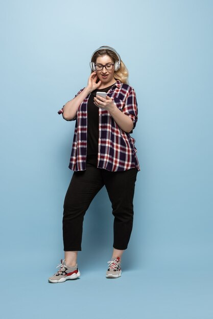 Mujer joven en ropa casual con smartphone y auriculares en la pared azul. Carácter positivo del cuerpo, feminismo, amarse a sí misma, concepto de belleza. Plus tamaño hermosa empresaria. Inclusión, diversidad.