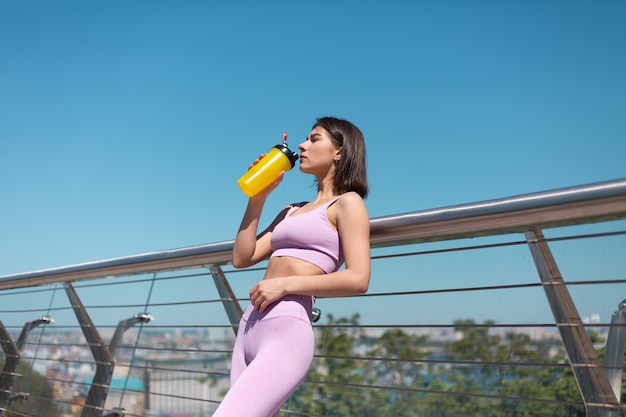 Mujer joven en ropa deportiva adecuada en el puente en la calurosa mañana soleada con una botella de agitador de agua sed después del entrenamiento cansado de beber