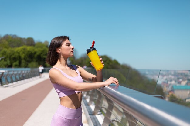 Mujer joven en ropa deportiva adecuada en el puente en la calurosa mañana soleada con una botella de agitador de agua sed después del entrenamiento cansado de beber