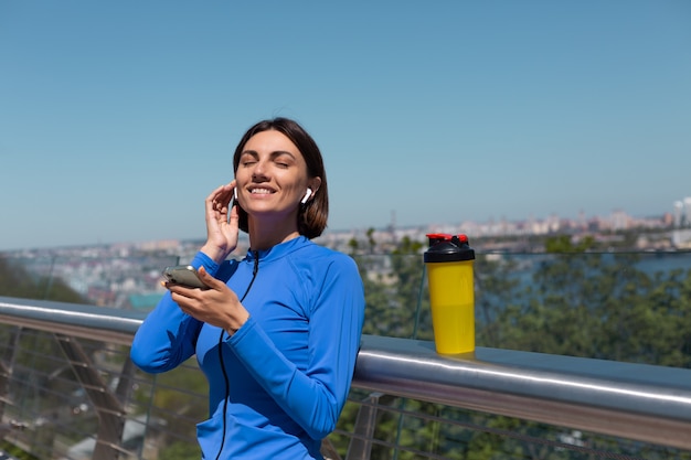 Mujer joven en ropa deportiva azul en el puente en la calurosa mañana soleada con auriculares inalámbricos y teléfono móvil, descansando escucha música