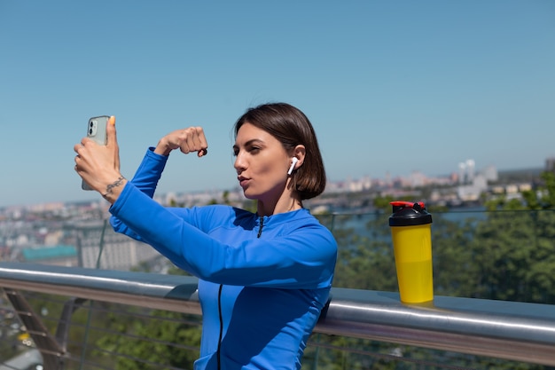 Mujer joven en ropa deportiva azul en el puente en la calurosa mañana soleada con auriculares inalámbricos y teléfono móvil, tomar selfie foto video para redes sociales muestra sus músculos bíceps