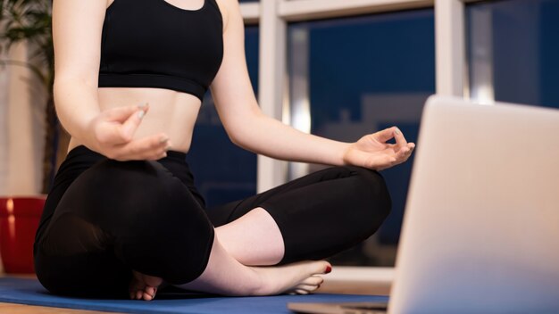 Mujer joven en ropa deportiva está meditando sobre una estera de yoga con un portátil delante de ella