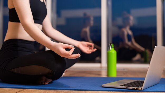 Mujer joven en ropa deportiva está meditando sobre una estera de yoga con un portátil delante de ella
