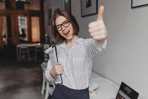 Foto gratuita mujer joven en ropa de estilo de oficina y gafas sostiene tableta con documentos, guiña un ojo y muestra el pulgar hacia arriba.