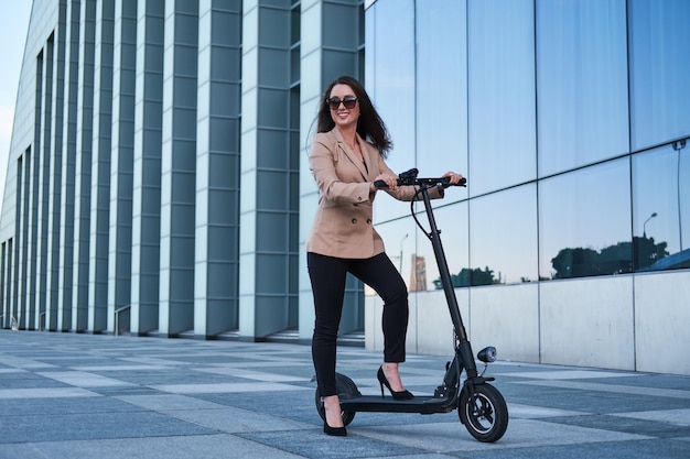 Mujer joven en ropa formal y anteojos está montando scooter eléctrico de su trabajo.
