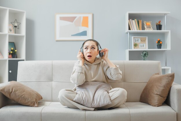 Mujer joven con ropa informal con auriculares sentada en un sofá en el interior de casa mirando confundida y preocupada