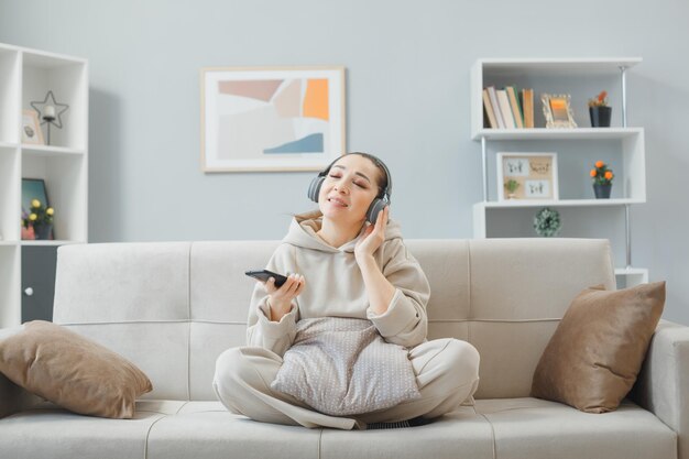 Mujer joven con ropa informal con auriculares sentada en un sofá en el interior de su casa usando un teléfono inteligente escuchando música disfrutando de su tiempo
