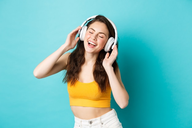 Mujer joven en ropa de verano escuchando música, usando audífonos y cantando su canción favorita, bailando en auriculares, de pie contra el fondo azul.