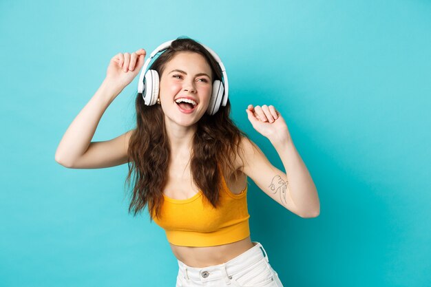 Mujer joven en ropa de verano escuchando música, usando audífonos y cantando su canción favorita, bailando en auriculares, de pie contra el fondo azul.