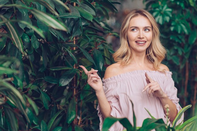 Mujer joven rubia contemplada que se coloca cerca de las plantas verdes que señalan su dedo