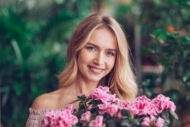 Mujer joven rubia feliz que se coloca detrás de las flores rosadas con el fondo borroso