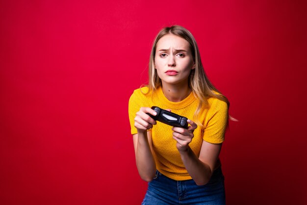 Mujer joven rubia gamer con gamepad jugando videojuegos aislados sobre pared roja