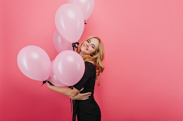 Mujer joven rubia posando con sonrisa feliz en la pared brillante. Foto interior de feliz cumpleañera europea sosteniendo globos.