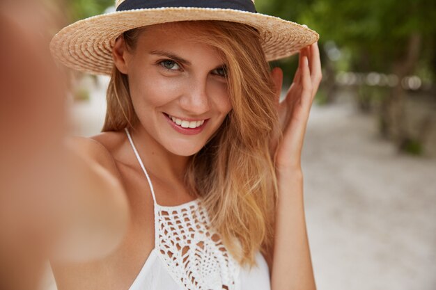 Mujer joven rubia positiva con expresión alegre hace selfie como poses al aire libre en una isla tropical, lleva sombrero de verano de moda y vestido blanco